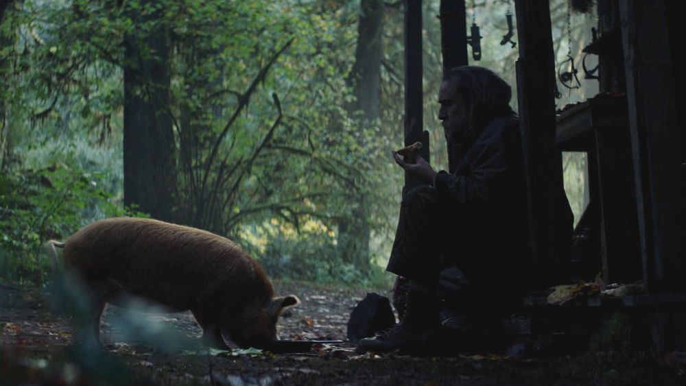 Nicolas Cage as Robin in Pig, sitting on his doorstep with his pig in front of him. She eats from a pan while he chews on a pastry.