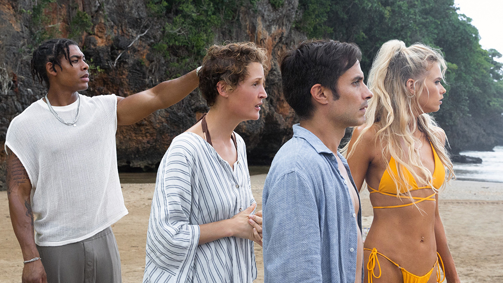 Aaron Pierre as Brendan, Vicky Krieps as Prisca, Gael García Bernal as Guy, and Abbey Lee as Chrystal in Old. The group stands on the beach, looking out at the water as Pierre points at something off-screen.
