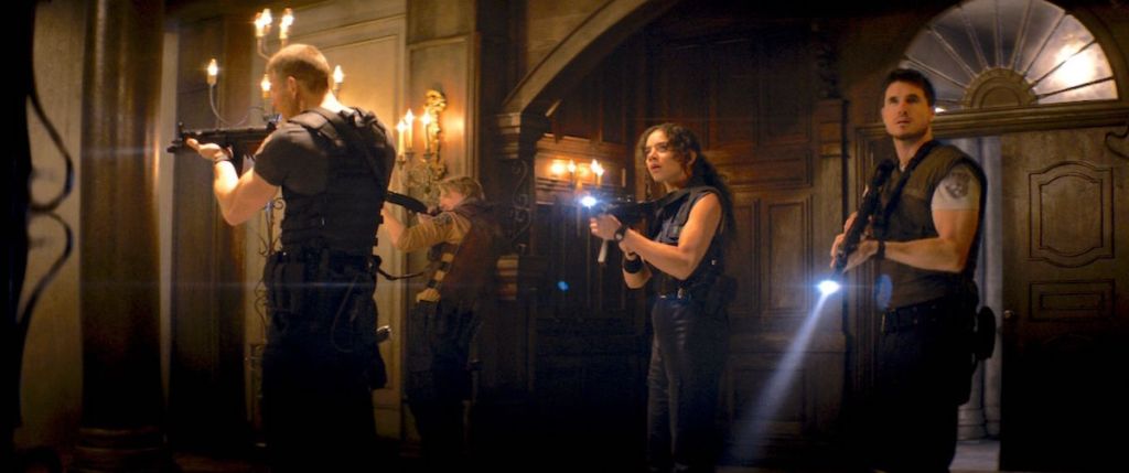 Wesker, Jill, and Chris stand in the lobby of the Spencer Mansion, flashlights on their guns pointing around the room