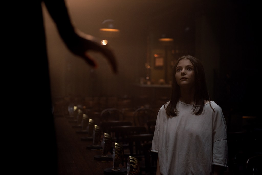A young girl with long dark hair (McKenzie) wears an oversized white t-shirt. She stands in an auditorium that appears empty, looking up at a woman performing, who is partially obscured by both the framing and the lighting.