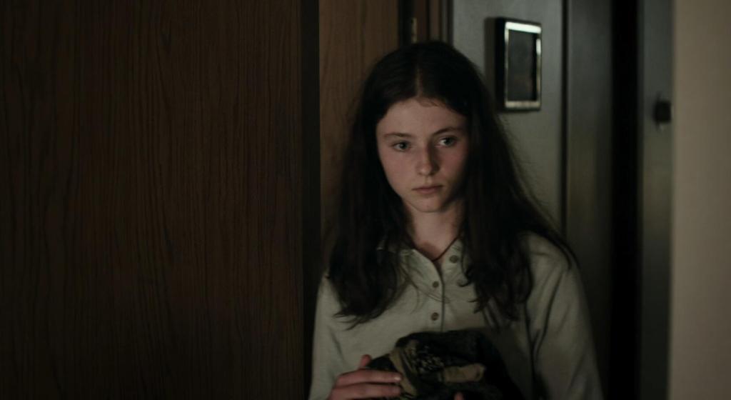 A young girl with long brunette hair (McKenzie) wearing a button-up shirt. She is inside a building, and holds clothing in her arms.