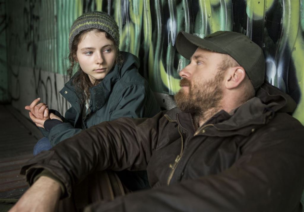 A young girl with brunette hair (McKenzie) wearing a patterned green beanie and green jacket. She sits against a graffitied wall with a middle-aged white man (Foster), who has a rugged beard, brown zip-up jacket, and green baseball cap. The two are looking at one another.