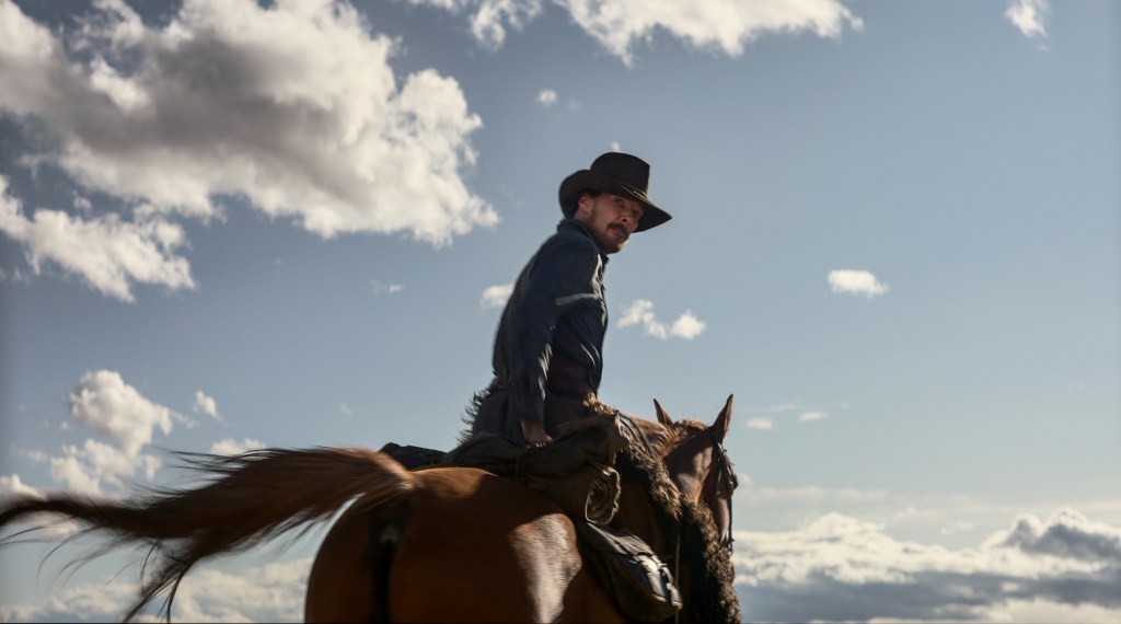 A man (Cumberbatch) wears a cowboy hat and shirt, looking over his shoulder while riding a brown horse. The horse is seen from behind, with a blue sky and clouds as the backdrop.