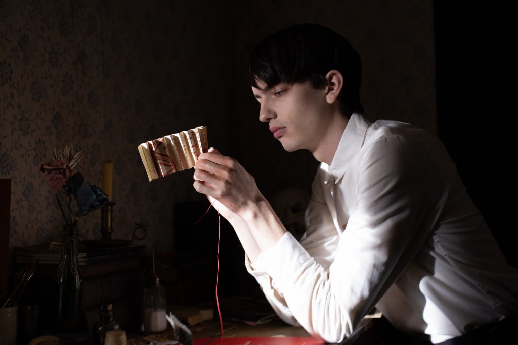 A young boy with short dark hair (Smit-McPhee) in a white shirt. He is seen looking at an object intently and using red thread while sitting at a table. Some of the thread can be seen dangling.