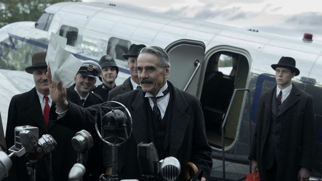Jeremy Irons' Neville Chamberlain stands in front of a set of microphones with a small aircraft behind him, having returned with the Munich Agreement that he is happily waving