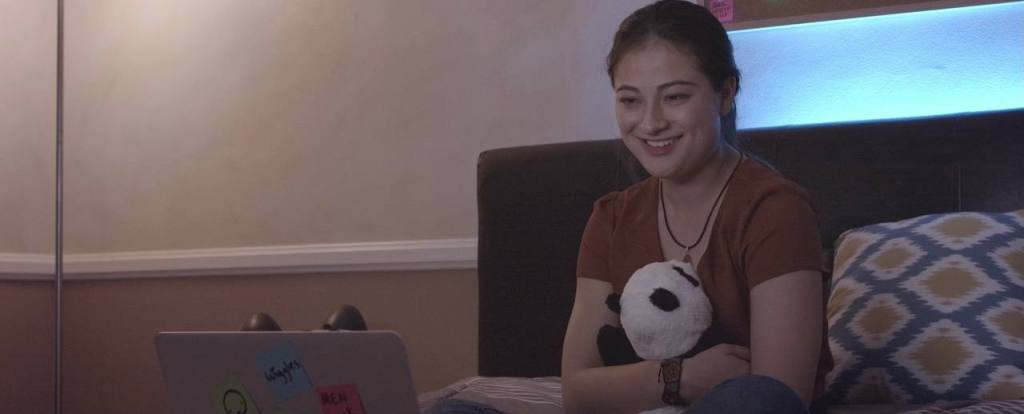 Nicole Ohara as Elyse is seated on her bed, hugging a panda plushie in her room while she FaceTimes her girlfriend using her laptop. She is smiling.