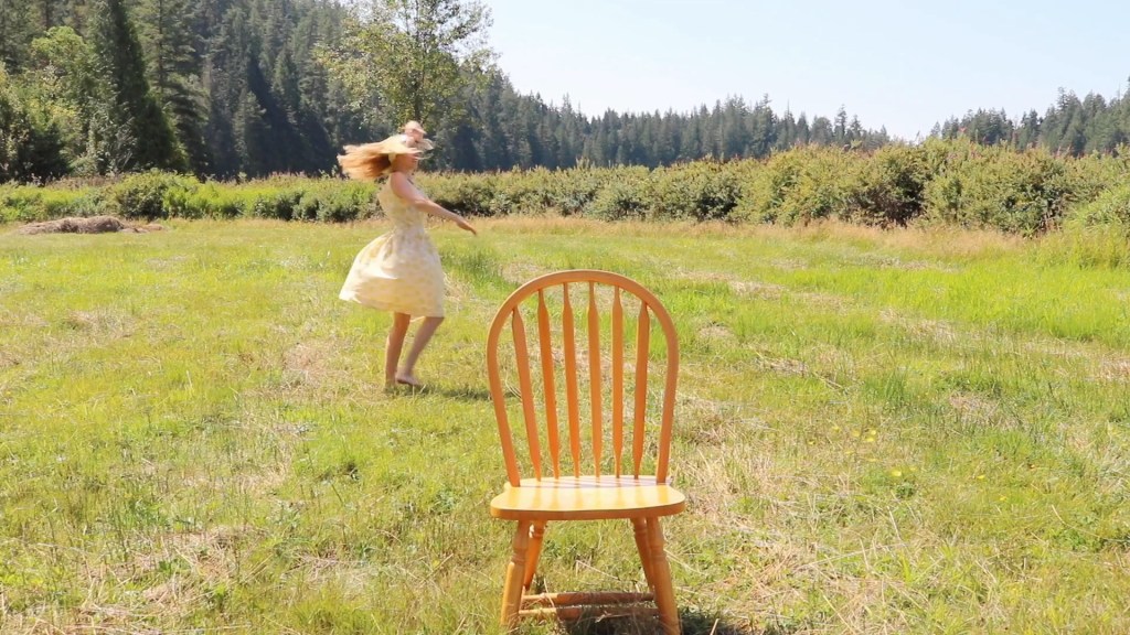A girl dances around in a grassy area while wearing a yellow dress. An empty chair is in the foreground of the shot.