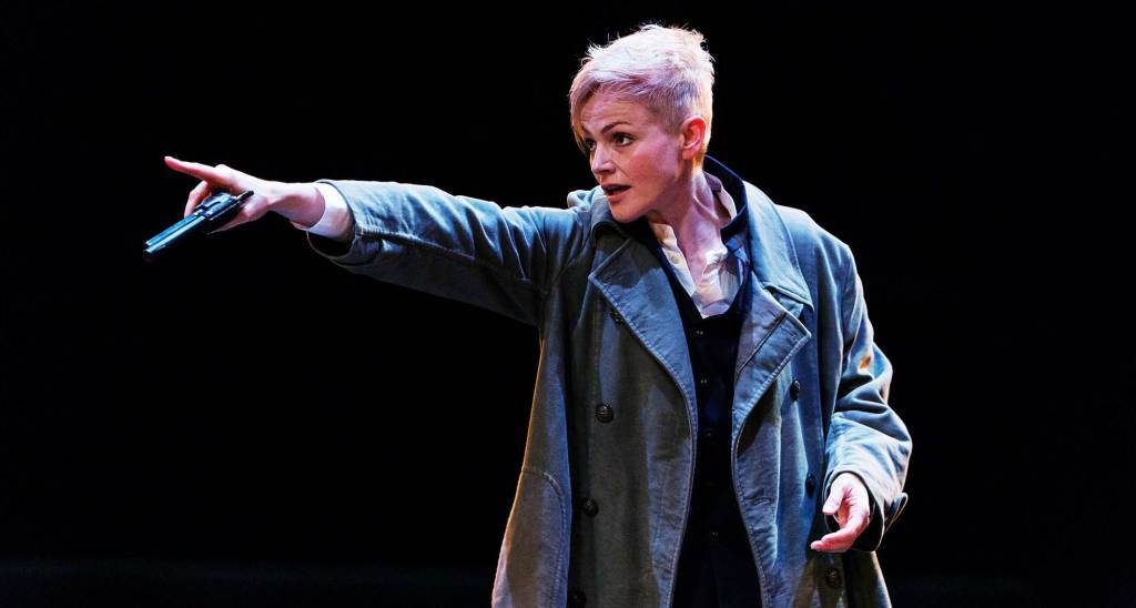Maxine Peake as Hamlet at the Royal Exchange Theatre. She is backdropped by a black background and is wearing a murky green coat. She has a pixie cut. In this image, she is pointing a gun with her right hand.