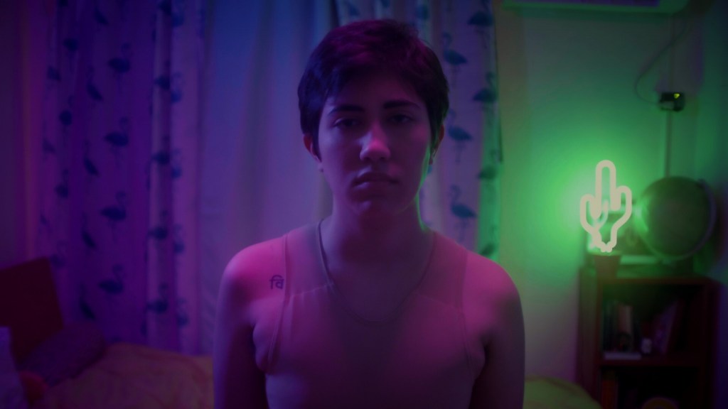 César stands in a darkened room, wearing only a binder on his upper half. He stares at the camera with a serious look on his face