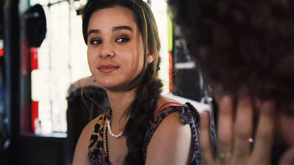 Violet Mulligan (Hailee Steinfeld) in Begin Again. She wears a leopard print tank top and a shell necklace, with her hair in a side braid. She is sitting at a table looking at a man whose face is not visible in the frame.