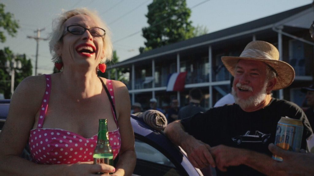 Tara is laughing with her head high int he air, wearing a polkadot dress and strawberry earrings. A man in a straw hat leans against a car chatting whilst another, his body just out of shot, holds a beer and listens in