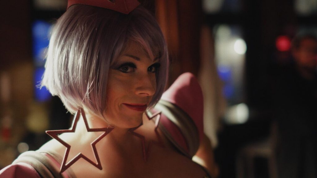 Tara looks sidelong at the camera, wearing a purple bob, a heavily shouldered outfit, and enormous star-shaped earrings