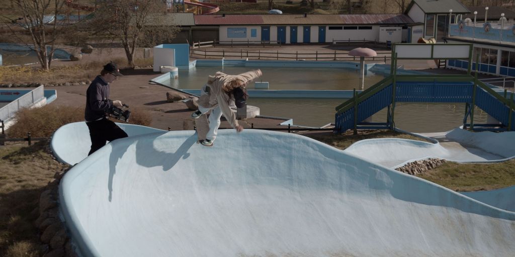 Still from 'Oski' showing Oski skating along the coping of a light blue water slide at a closed off and empty water park. Close to him, to the left, is his friend Tor Ström filming him. 
