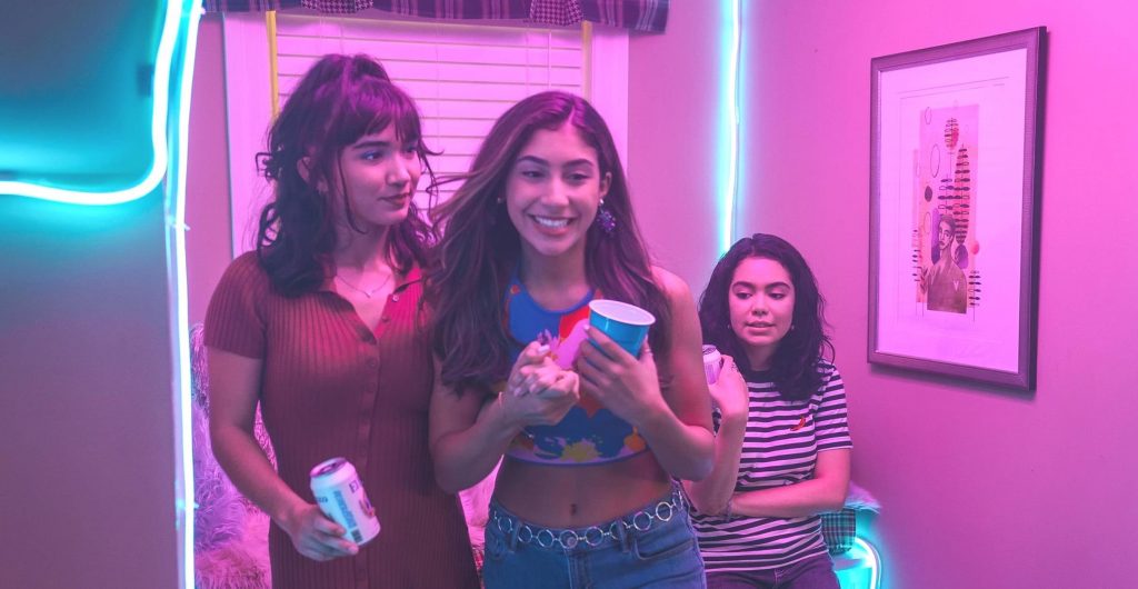A still from 'Crush' (2022) featuring three girls in a room awash in colourful pink light. The girl on the left has her hair done up and wears a red dress, looking to her right. The girl in the middle has long brunette hair and wears a patterned crop top and jeans while smiling. The girl on the right has mid-length dark wavy hair, and she wears a striped shirt and appears smaller in the frame.