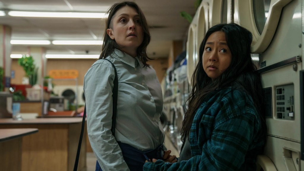 A still from 'Everything Everywhere All at Once' of two girls at a laundromat. The girl on the left is taller and has an undercut, wearing a collared shirt with a bag slung over her shoulder. The girl on the right is shorter and wears a green and blue plaid shirt. They both have concerned expressions.