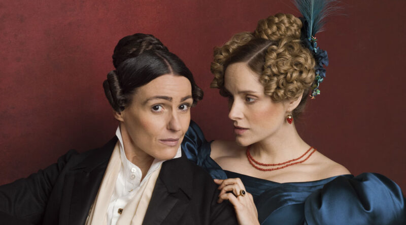A promotional still for 'Gentleman Jack' featuring Suranne Jones as Anne Lister and Sophie Rundle as Anne Walker. Jones wears a black suit with her hair done up, while Rundle wears a poofy blue dress with her hair in a tight curly updo, accessorizing with a blue feathered hat. She looks at Jones, who is looking at the camera.