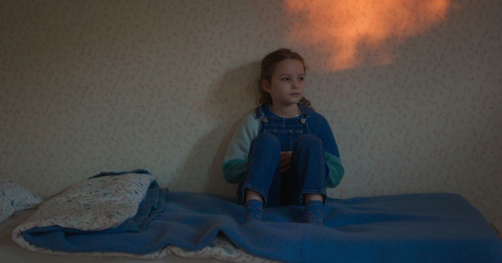 Nelly (Josephine Sanz) sitting on her bed. She is against the wall, wearing overalls and a hoodie.