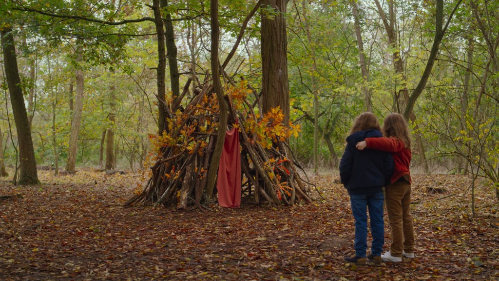 Nelly (Josephine Sanz) and Child Marion (Gabrielle Sanz) seen from behind standing in a forest in front of a leaf-covered tent. Nelly is on the left wearing a dark sweater and jeans, while Child Marion wears a red sweater and brown pants and puts her arm around her.
