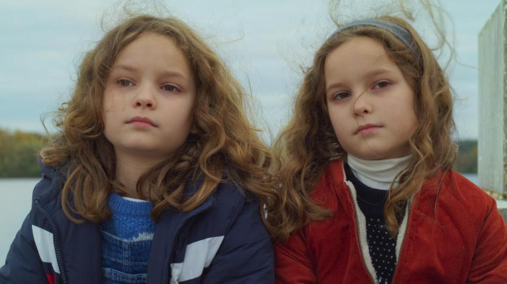Nelly (Josephine Sanz) and Child Marion (Gabrielle Sanz) sitting next to one another, facing the camera. Nelly wears a navy coat while Child Marion is in red. They have neutral expressions, and their wavy hair is windblown.