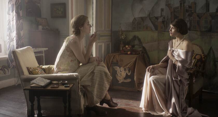 Elizabeth Debicki and Gemma Arterton as Virginia Woolf and Vita Sackville-West in a still from 'Vita & Virginia'. The two women sit across from one another, with Debicki wearing a flowing dress and sitting in a pensive pose as she studies Arterton. Arterton wears a shoulderless silky gown, facing Debicki.