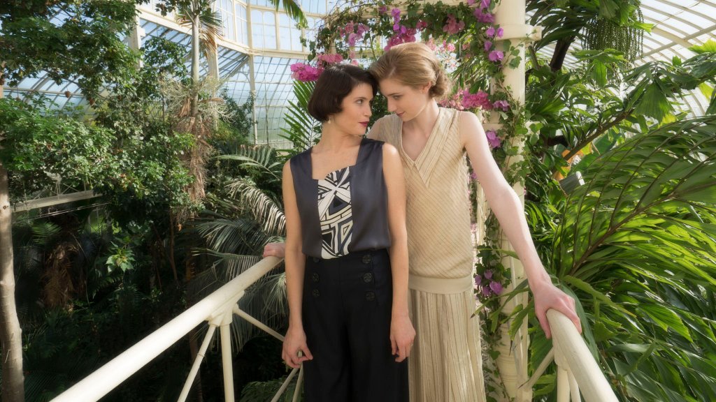 Gemma Arterton and Elizabeth Debicki as Vita Sackville-West and Virginia Woolf in a still from 'Vita & Virginia'. They are in a greenhouse, with Arterton wearing a vest, top, and trousers, as Debicki stands tall above her in a long yellow dress. The two are surrounded by plants.