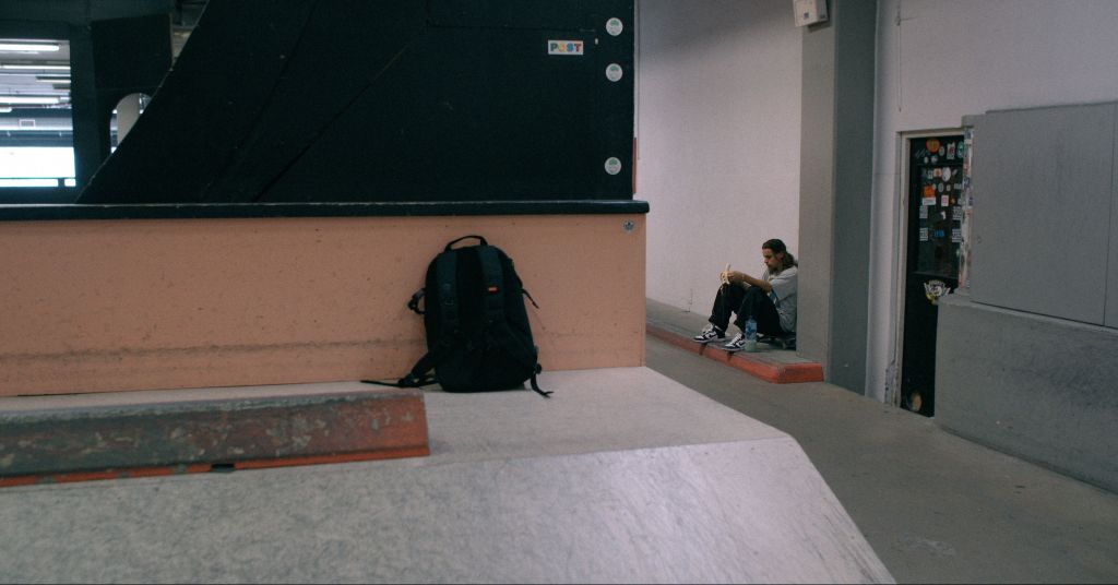 Still from 'Oski' showing Oski sitting alone in the distance, leaning against a white wall while sitting on a curb inside of an indoor skatepark. He's eating a banana and looking down at the ground.