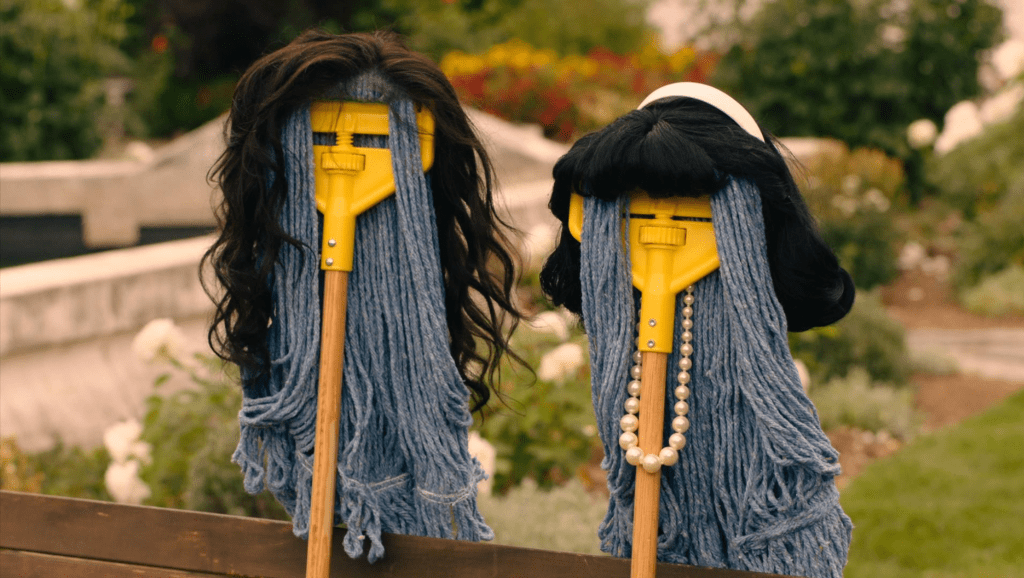 A close-up still of two frilly mops sitting on a bench outside in a park. They are supposed to resemble two Debras, Jouhari (there's a dark brown wavy wig attached to the top of the mop) and Honig (there's a black wig with a fringe and a white headband attached to the top of the mop as well as a pearl necklace). 