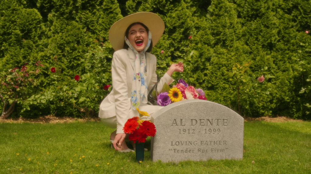 Still of Stonoha's Debra squatting down behind a gravestone with a huge smile on her face. She's wearing all white, a big hat and carrying a basket of perfectly looking flowers. She's just about to continuing flower-picking, by taking the flowers next to the gravestone she's behind. The gravestone says: "AL DENTE, 1912-1999, Loving Father, 'Tender But Firm'"