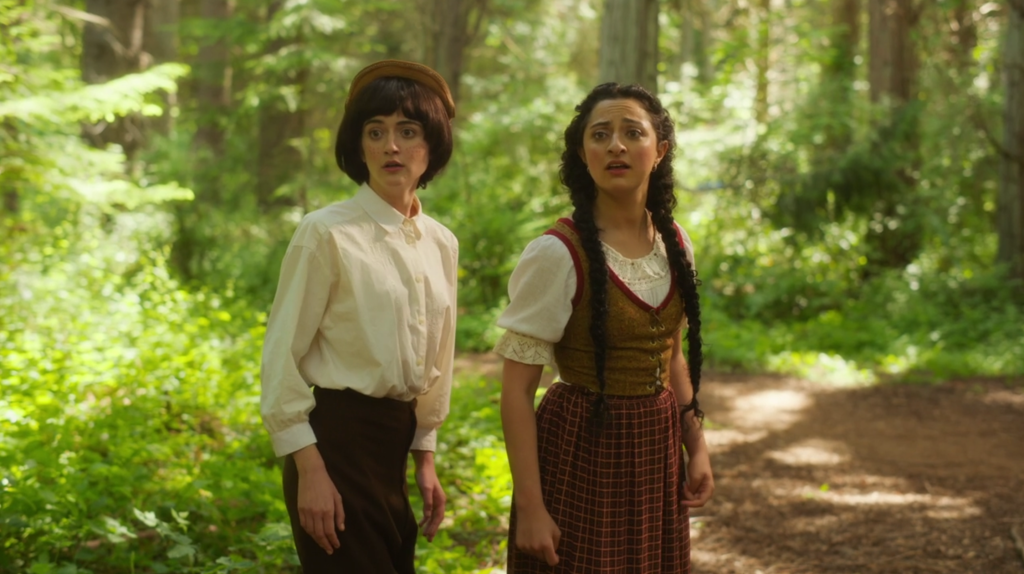 Still showing two Debras in the forest dressed in Hansel (Stonoha) and Gretel (Jouhari) costumes during an adventure to save the remaining Debra (Honig). They both have surprised expressions on their faces, as if they've just turned around and noticed something in the distance behind them. They're looking towards something out of frame.