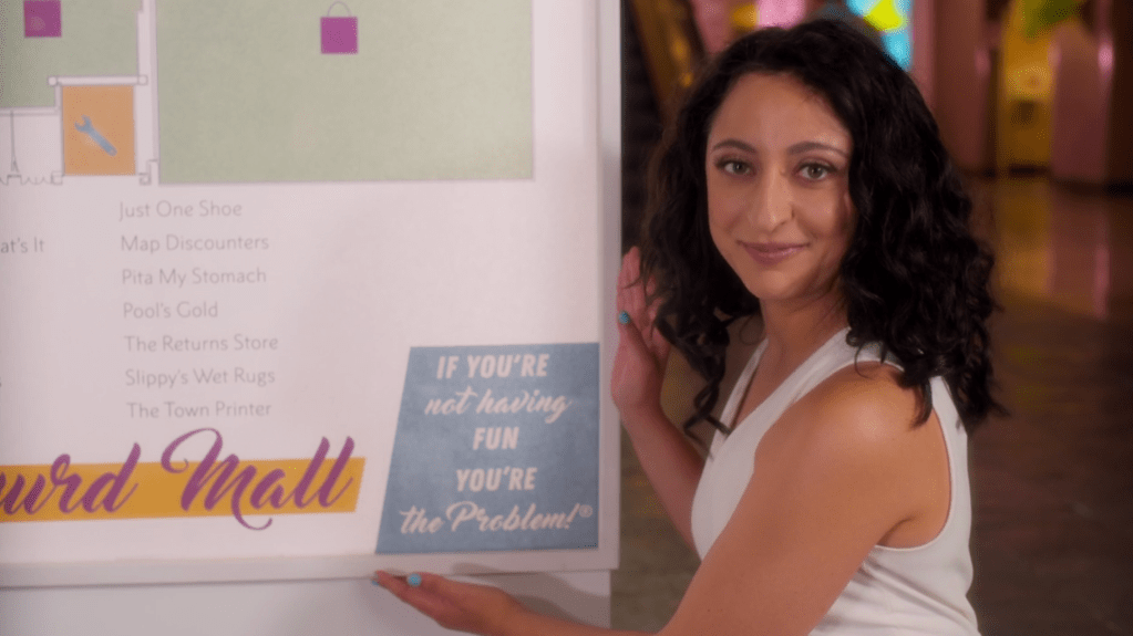 A still of Debra (Jouhari) looking directly into the camera, while smiling and positioning her hands to emphasis the slogan "If you're not having fun you're the problem" on a sign from the local mall. Next to the slogan, various names of stores located inside the mall are seen, for example: Just One Shoe, Pita My Stomach, The Returns Store and Slippy's Wet Rugs.