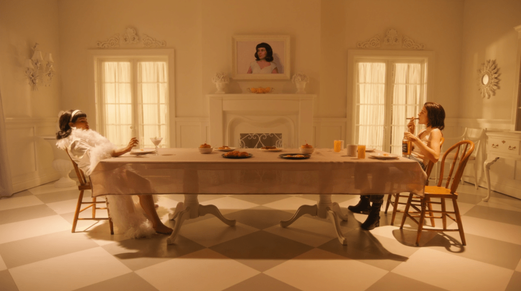 A still showing Debra's (Honig) white living room where all brunches are held. In this scenario, Honig and Stonoha sits at the opposite end of the dinner table. To the left is Honig, dressed in a white frilly dressing-gown, and to the right is Stonoha, dressed in black boots, blue jeans, a white tank top as she's smoking on a cigar and holding a bottle of whiskey. They're intensely looking at each other.  