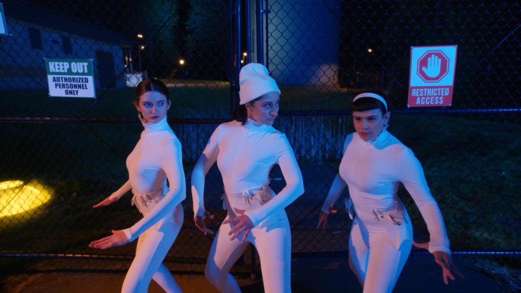 Still showing the three Debras before breaking into the restricted area to steal Lemoncurd's milk supply. They're standing in front of a fence (with various visible "KEEP OUT" signs) in all white and tight outfits resembling what a Charlie's Angel might wear to a mission. They each have a pose which looks very staged and not very natural as they look suspiciously around while their arms are positioned mechanically from their bodies. 