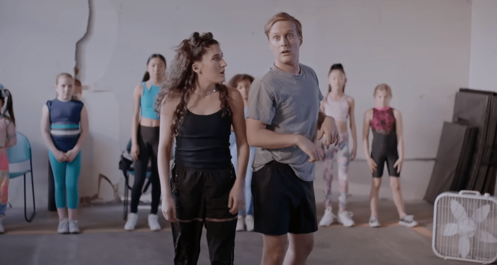 Still from the dance class sketch. In the background several young girls are seen standing in a line against the wall, all wearing colourful athletic wear. In the foreground, centered in the frame, is Berlant and Early, also wearing athletic wear but in black and grey tones. Berlant, who has her curly hair out except for two smaller space buns in the front, is looking at Early with an open mouth and flabbergasted expression. Early is looking towards the dance teacher (outside of frame) with a confident facial expression and wide open eyes as if he just said something to challenge her. 