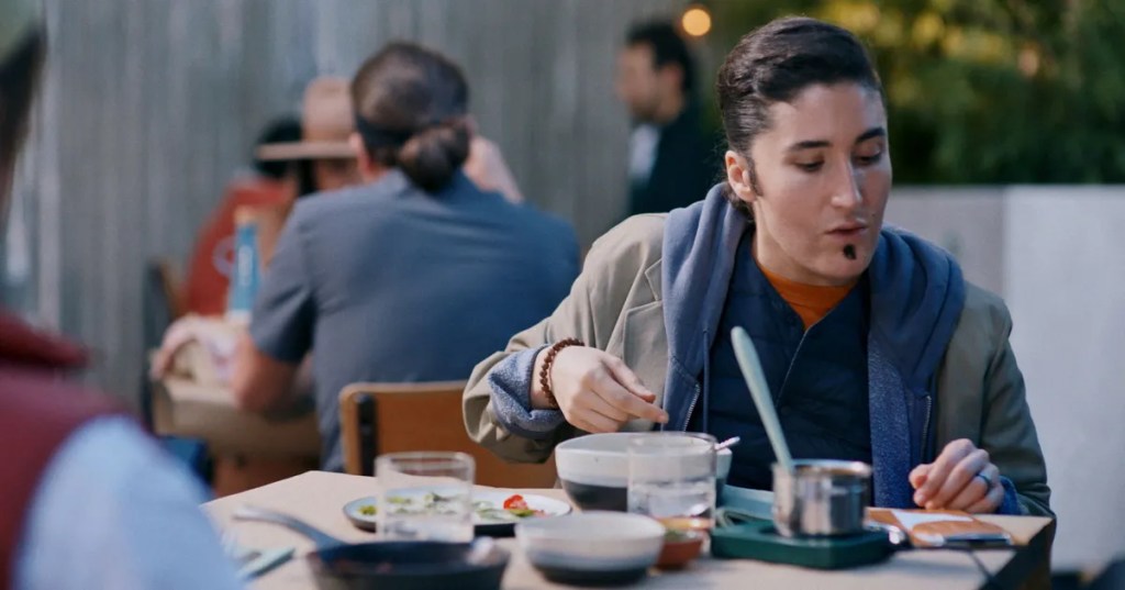 Still from one of the hot caramel sketches. Pictured is Berlant sitting down at a table at a restaurant while dressed like a man, complete with her hair slicked back and fake facial hair. Behind her is other restaurant goers and in front of her barely outside of the frame is Early. On their table various empty and half-empty dishes, bowls and glasses are seen. Berlant has her hot plate in front of her and has poured hot liquid onto the paper bill in front of her. She's currently looking at the bill, as she's trying to calculate how much tip to leave. 