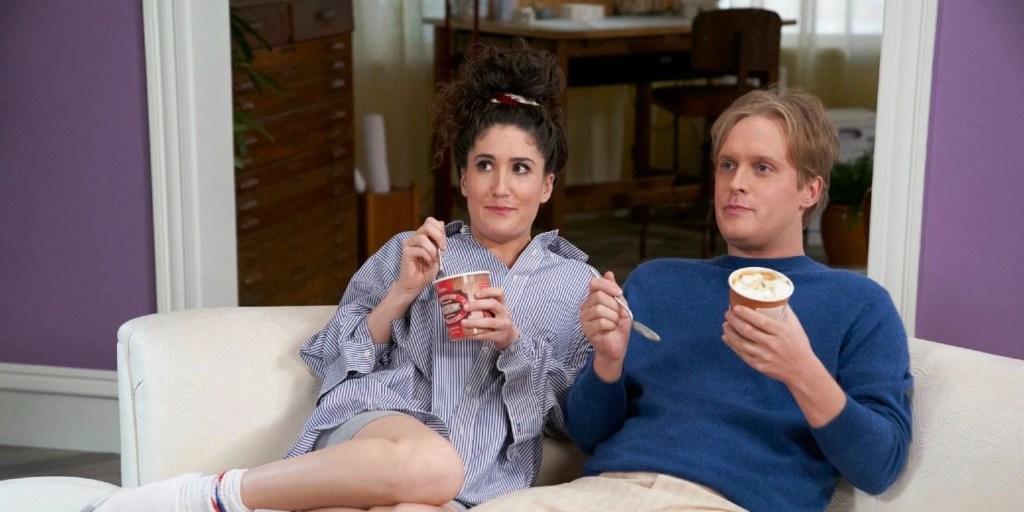 Still from the fictional sitcom "He's Gay, She's Half-Jewish." It shows the two characters portrayed by Berlant and Early sitting comfortably on a white sofa in their apartment, looking towards something outside of the frame. Berlant is wearing an oversized button-up shirt, grey shorts and white socks (laidback, comfortable and casual) while Early is wearing light beige pants and a blue knitted sweater (formal). They're both eating ice cream, directly from the tub. They have one each as well as a spoon each. Berlant looks curiously ahead with wide eyes, while Early looks neutral and borderline bored and tired. 
