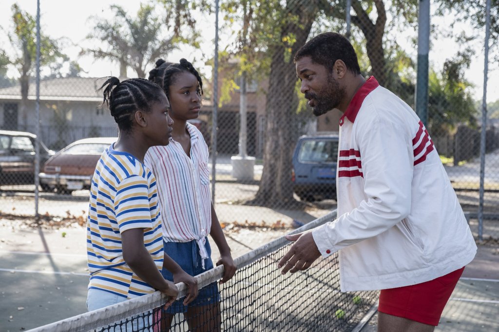 A still of Will Smith, Saniyya Sidney, and Demi Singleton in 'King Richard'. They stand on a tennis court, with Sidney and Singleton both wearing striped shirts and standing behind the net while Smith speaks to them.