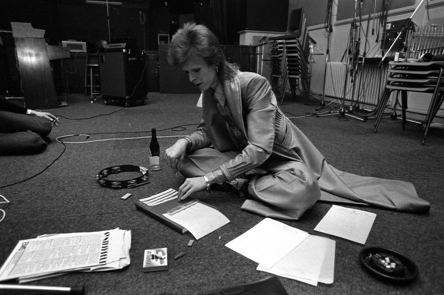 Bowie in black and white, doing the cut up method of songwriting, in a music studio. Next to him is a tambourine, beer bottle and ashtray. He wears a long leather coat and flowing trousers. 