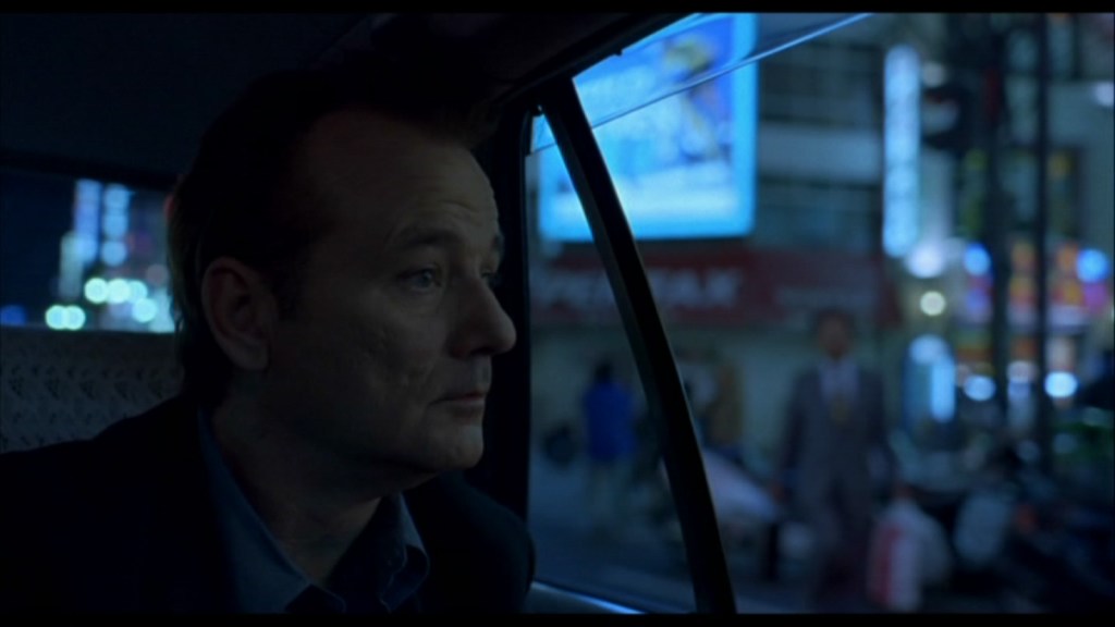 A still of Bill Murray as Bob in 'Lost in Translation'. He is seen from inside a car, looking out the window with the streets of Japan a blur in the exterior.