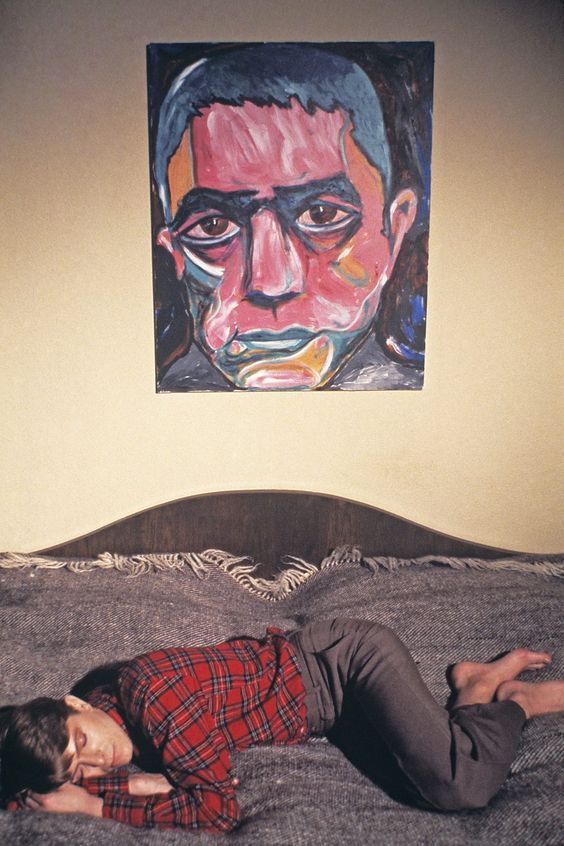 David Bowie lies on a grey bed barefoot, with a tartan red shirt and grey trouers. On the wall huge above him, is his portrait of a man with a red and orange face, with furrowed brows and big eyes. He is Yukio Mishima, a Japanese author. 