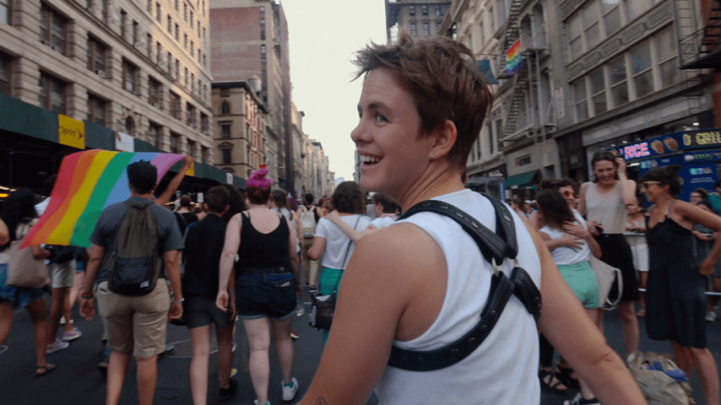 Taking part in New York City's Pride March, Leo Baker is shown from behind, lightly turning his head to smile towards someone outside the frame. He's wearing a white tank top, a black harness and a short haircut. Out of focus in front of him is many people and buildings seen, as well as several rainbow flags.