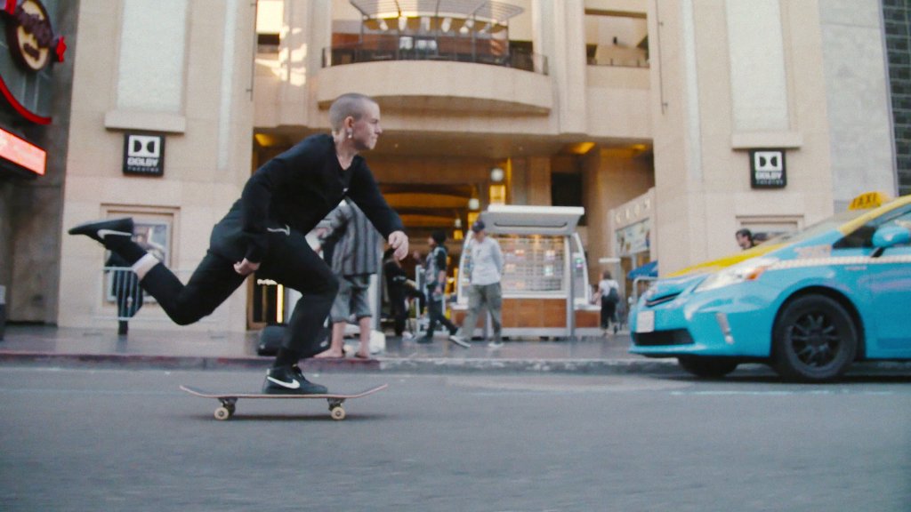 Leo Baker shown on his skateboard, pushing down a street in New York City. He's wearing all black and sports a buzz cut. He's caught in motion, right foot firmly on his board as his left one is in the air after pushing it into the ground to gain speed.