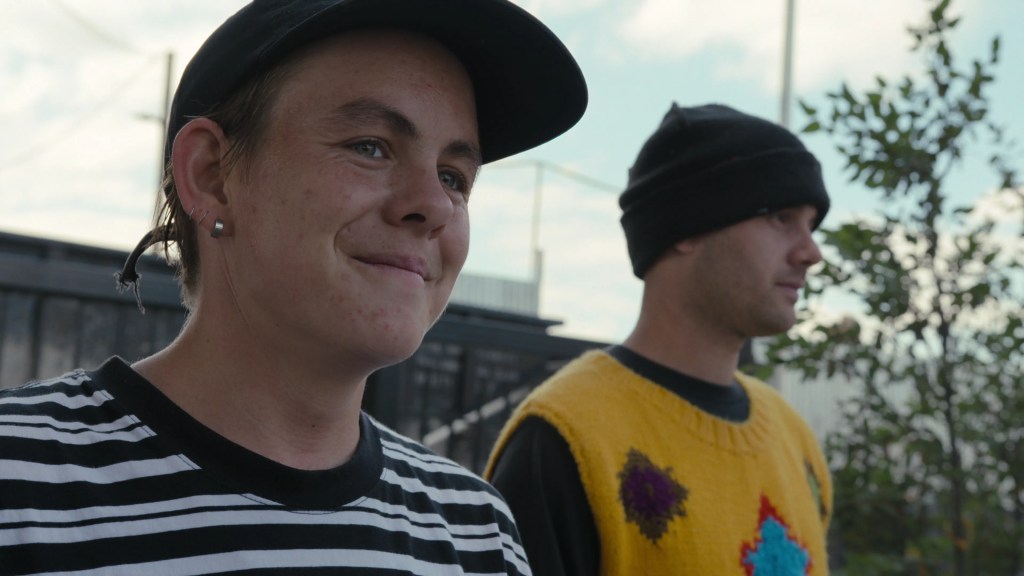 Baker shown in focus in a close-up, smiling towards someone outside of frame. He's wearing a black cap and a black-and-white striped shirt. Beside him out of focus is Stephen Ostrowski, wearing a black beanie, a black shirt and a yellow knitted vest. They're outside, with blue skies, clouds and trees are visible. 