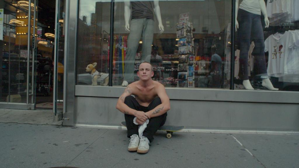 Baker seen sitting outside in front of a display window of a store. He is wearing lightly coloured shoes, black pants and is holding his white shirt in his hands as his naked upper body is shown. He is sitting on his skateboard looking ahead with a neutral facial expression. 