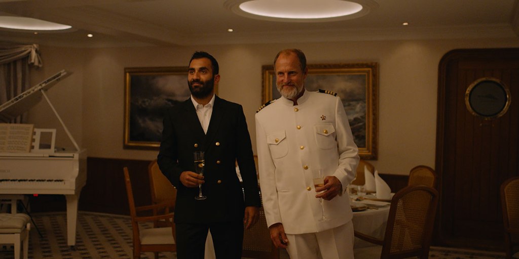 Two men standing in an extravagant dining room on a cruise ship. The man on the left has a beard and wears a suit, while the man on the right wears a white captain's outfit. Both are seen smiling and holding drink glasses.