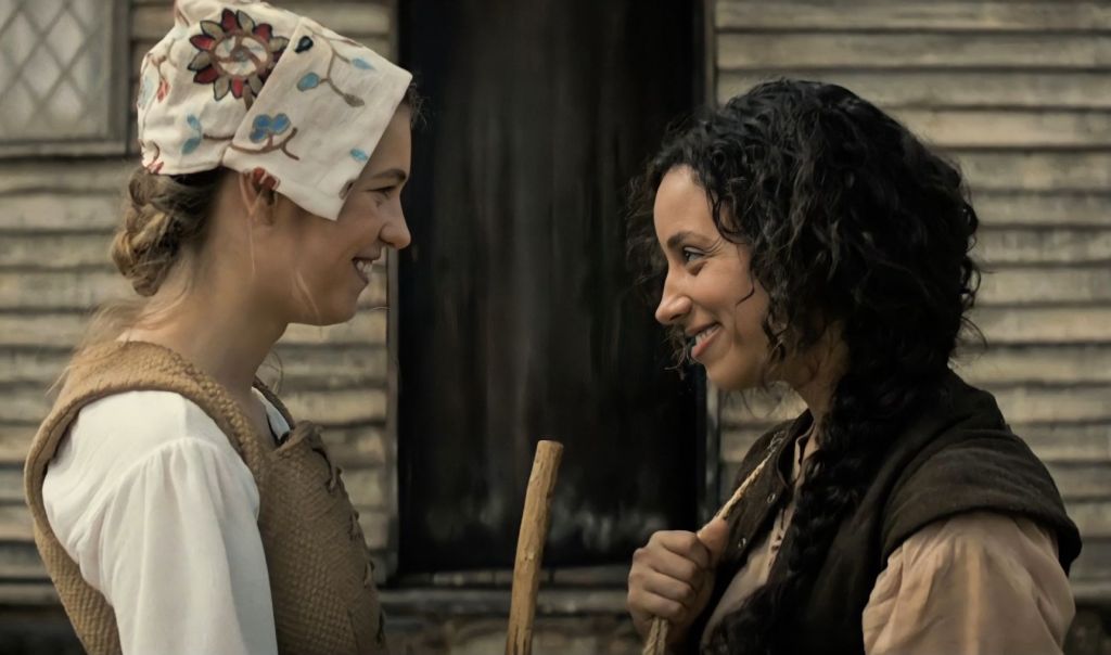 A still from 'Fear Street Part 3: 1666'. Hannah (Olivia Scott Welch) and Sarah (Kiana Madeira) are seen in profile view smiling at one another, dressed in period clothes.