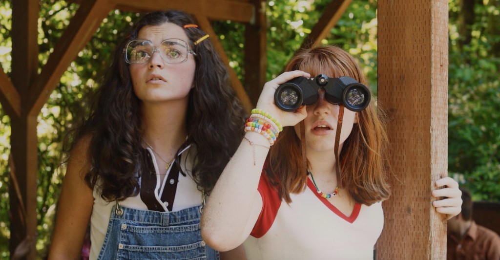 A still from Camp Calypso showing two young women styled in 1970s clothing looking ahead towards something outside of the frame. One of them is using binoculars. 