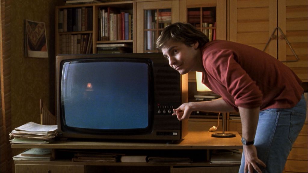 Alex (Daniel Brühl) in Good Bye, Lenin! looking towards someone outside of frame, whilst trying to find a channel on a nearby television by touching a button. He's wearing light blue jeans, a watch with a black strap, and a red jumper. Behind him and the television are bookshelves.