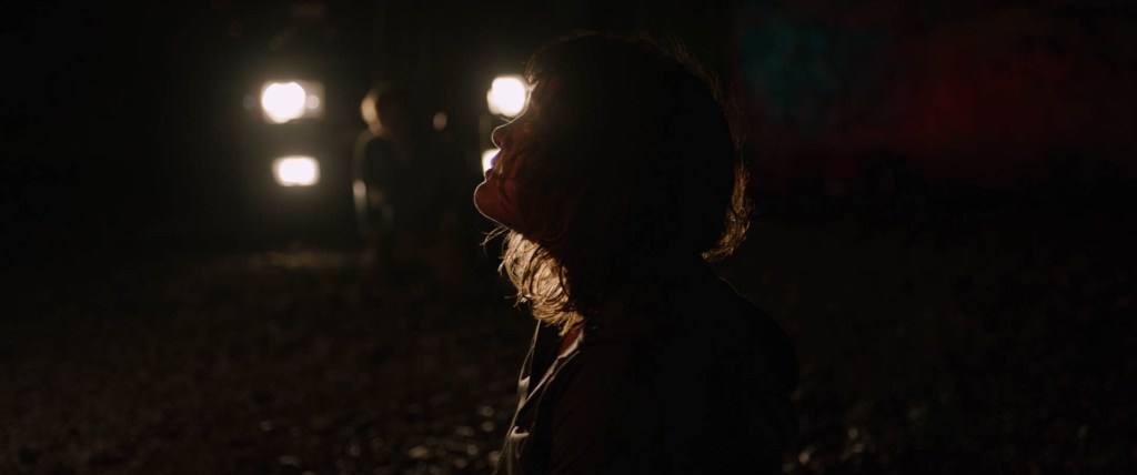 Still showing Jill from her profile as she's on her knees on the ground. She's looking up towards someone outside of the frame, with a pleading facial expression. Behind her, one can see the strong headlights from the car used for the kidnapping shining as well as Hoaggie on his knees in front of the car between the light sources, barely seen since it's so dark besides the headlights. 