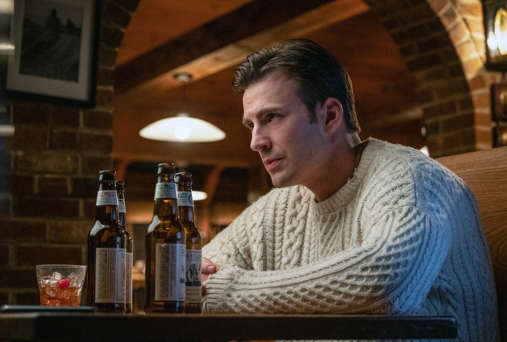 Ransom (Chris Evans) from Knives Out sitting in a booth at a bar looking at Martha (Ana de Armas) outside of frame. In front of him are four empty bottles of beer and a newly ordered drink. He's wearing the white knitted jumper, with its tears and holes clearly visible.