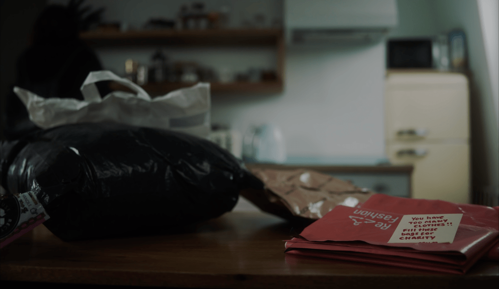 A still from Material Girl showing several plastic bags containing new clothing items scattered on a table in a kitchen. Next to them are a bunch of plastic bags used to donate clothing items with a post-it note on top saying: "You have too many clothes!! Fill these bags for charity." 